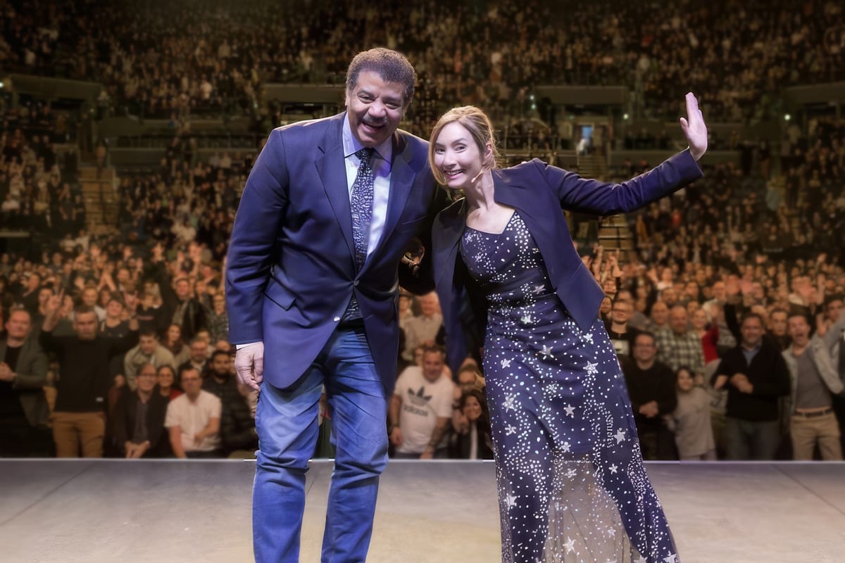 Dr Michelle Dickinson on stage with Neil deGrasse Tyson, waving to a packed arena audience